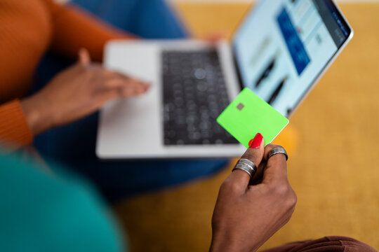 A close-up of a woman's hand holding a bright green credit card while shopping online on a laptop, with a blurred screen displaying an e-commerce website.