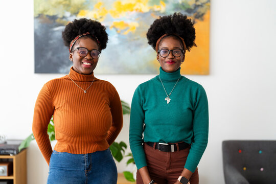 Two young women with natural curly hair, wearing glasses and turtleneck sweaters, standing side by side in a modern indoor setting with a colorful abstract painting in the background.