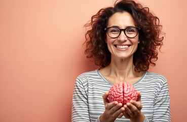 Middle-aged brunette woman smiling confidently holds brain model against pink background. Happy lady with eyeglasses shows white teeth, promoting mental health care, memory enhancement, cognitive