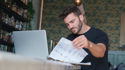 Male Owner Or Manager Of Restaurant Going Through Paperwork With Laptop At Bar