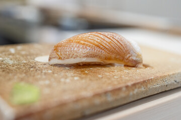 Close-up of a sea bream fry nigiri sushi on a textured cutting board, highlighting the delicate texture and precise knife cuts. The glossy fish surface reflects the light, enhancing its freshness
