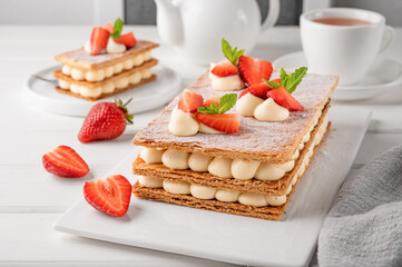 French dessert millefeuille with vanilla cream and fresh strawberries on a white plate on a white wooden background.