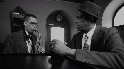 Black and white shot of saxophonist in checkered suit playing soulfully while Black man in hat listening intently across piano in vintage themed jazz club