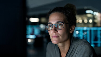 Obraz premium Thoughtful female IT specialist working late at night in a modern tech office, screen light reflecting in her glasses, focused on digital data – Generative AI