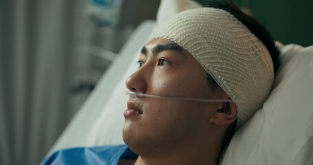 A young man with a bandaged head and an oxygen tube rests in a hospital bed, gazing through the window as he recovers under medical supervision in a quiet and well-equipped ward.