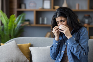 Indian young woman sitting on sofa at home and wiping nose with tissue, suffering from runny nose...
