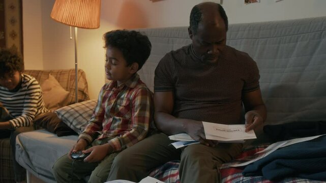 African American man hugging his younger son, while talking to his brother and looking through bill, sitting on couch