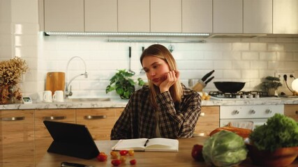 Beautiful girl eating apple watching podcast video on tablet sitting at table in kitchen of her house apartment, recipes, cooking, studying, student, eating, watching media, technology, display - Powered by Adobe