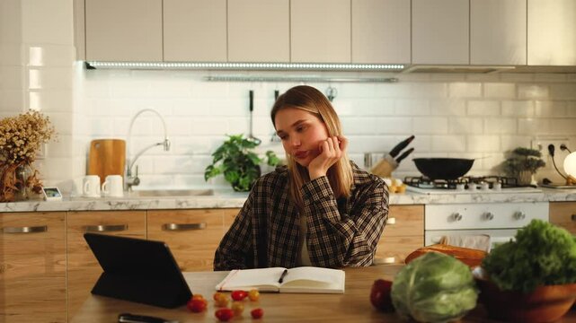 Beautiful girl eating apple watching podcast video on tablet sitting at table in kitchen of her house apartment, recipes, cooking, studying, student, eating, watching media, technology, display