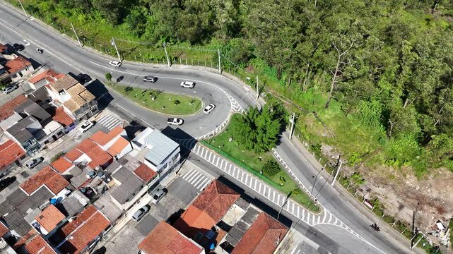 Vis&atilde;o a&eacute;rea de uma rotat&oacute;ria de tr&acirc;nsito em um dia ensolarado, com carros em movimento fluindo pelas ruas, capturada por drone, destacando a din&acirc;mica do tr&aacute;fego urbano e a infraestrutura rodovi&aacute;ria