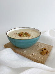 Creamy oatmeal porridge in a teal bowl on a wooden board on a white background. Healthy food with walnuts, kiwi, and oat flakes. Nutritious breakfast morning meal.