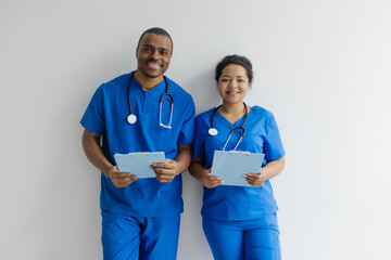 Close-up portrait of smiling African American male and female doctors in blue scrubs, friendly medical professionals standing with clipboards and stethoscopes, posing against white wall background