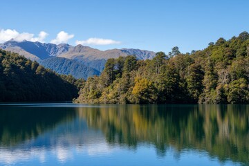 Fototapeta premium Serene lake reflects trees and mountains under a sunny, blue sky