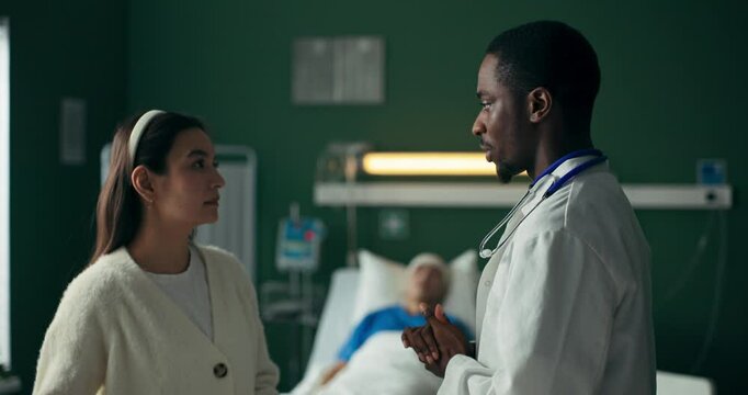 Young African doctor speaks with a woman, the hospital room behind them housing a critically ill patient.