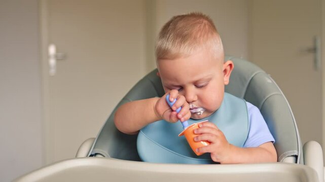 A one-year-old boy sits at a table and eats milk yogurt on his own. The child eats baby yogurt. The boy eats yogurt deliciously and smears it on his face.
