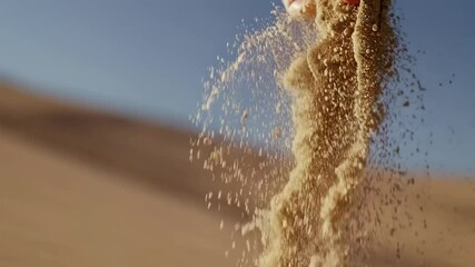 Close-up video of a hand releasing sand against a clear blue sky, shot from a low angle, capturing the flow and texture of the grains.