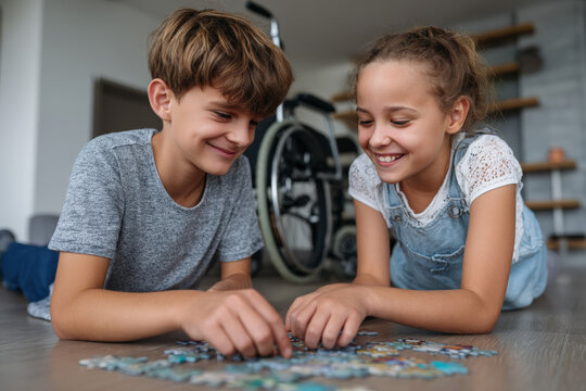 Two children, a boy and a girl, are happily assembling a jigsaw puzzle on the floor. The boy is around 12 years old, and the girl is about 10 years old. A wheelchair is visible in the background.