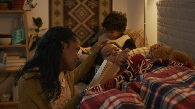 Loving African American mother reading her youngest son bedtime story, while her older son reading book by himself, sitting in armchair