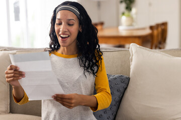 Excited woman reading letter on couch at home, celebrating good news