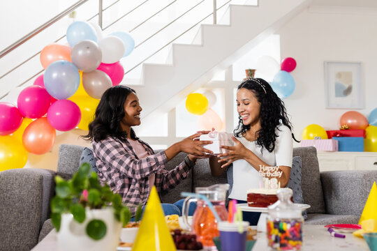 At home, celebrating birthday, diverse female friends exchanging gift on sofa with colorful balloons