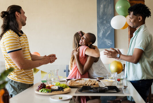 Diverse friends celebrating at home, hugging and clapping around kitchen island with snacks - Powered by Adobe