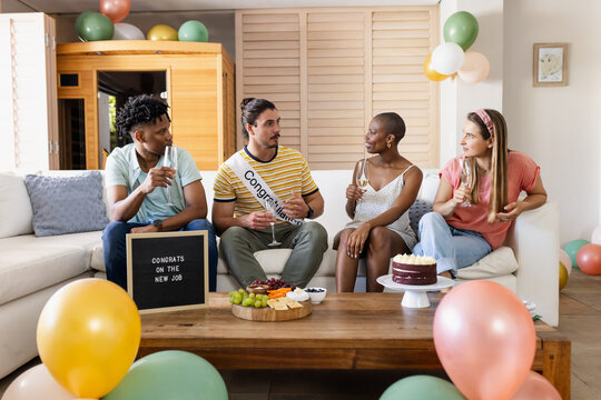 Diverse friends celebrating new job with drinks and snacks at home, feeling joyful - Powered by Adobe