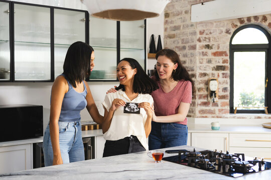 Celebrating pregnancy ultrasound, diverse female friends sharing joyful moment in kitchen, at home