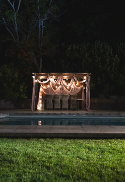 Outdoor pergola beautifully decorated with drapes and lights by pool at night