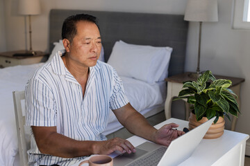 Senior Asian man using laptop at home, concentrating on work in bedroom