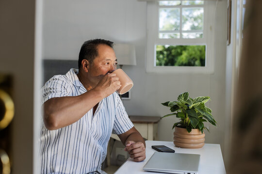 Senior Asian man enjoying coffee while working on laptop at home office desk - Powered by Adobe