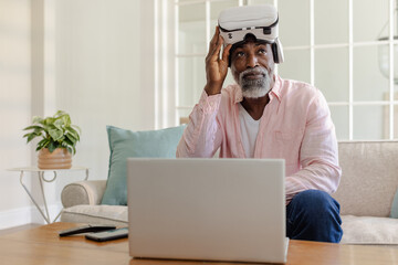 Senior man using VR headset and laptop at home, exploring virtual reality