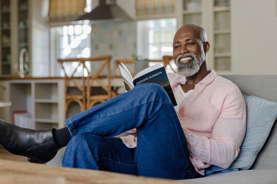 Smiling senior man reading book on sofa at home, enjoying leisure time