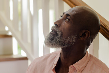 Senior African American man looking up thoughtfully while standing by staircase at home