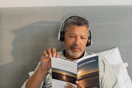 Senior man relaxing in bed, reading book and listening to music with headphones