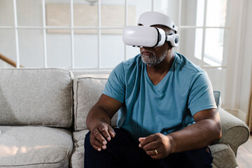 Senior African American man using VR headset sitting on sofa, experiencing virtual reality