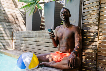 Relaxing by pool, African American man in orange shorts using smartphone, enjoying leisure time