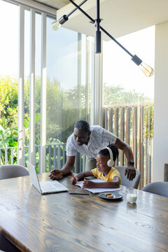 Father helping son with homework at home, using laptop and headphones
