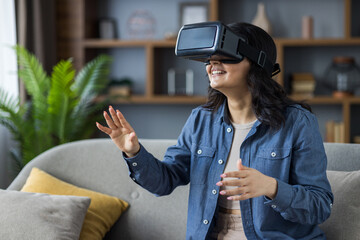 Indian young smiling girl sitting on sofa at home wearing virtual reality glasses and gesturing...