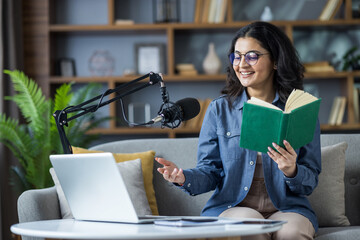 Smiling young Indian woman sitting on sofa at home, holding a book, reading and talking on laptop...
