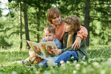 Fototapeta premium Candid scene of mother and sons reading book sitting together on picnic blanket in green Summer park copy space