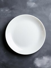 Close-up of a white plate on a dark grey textured surface. the plate is empty and appears to be made of ceramic or porcelain. it has a smooth, glossy finish and is slightly curved at the top.