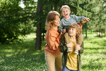 Fototapeta premium Waist up portrait of young boy holding little brother on shoulders with mother helping family enjoying time together in green park copy space 