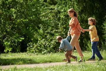 Obraz premium Full length side view of carefree mother with two boys holding hands and walking on path in green forest together lit by sunlight copy space
