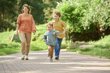 Fototapeta premium Full length front view of two brothers running towards camera on path in green park with mother walking in background copy space