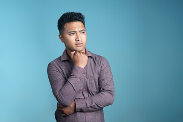 Fototapeta premium Young man in a maroon shirt looking away thoughtfully with hand on chin against a plain blue background. Concept of thinking, decision-making, and contemplation in studio setting.