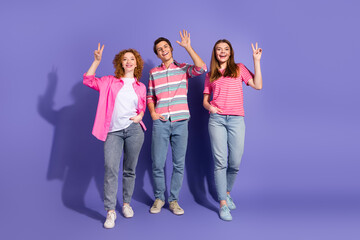 Fototapeta premium Group of three young smiling friends showing peace gestures on a vibrant purple background, promoting cheerful leisure