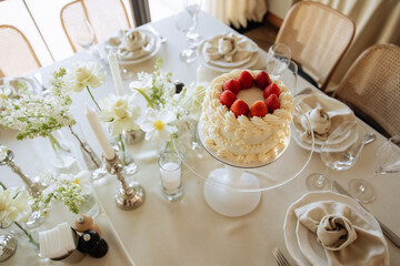 Elegant white cake with strawberries on top, placed on a glass stand, surrounded by flowers and candles on a table.