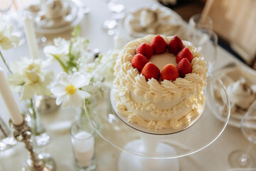Elegant white cake with strawberries on top, placed on a glass stand, surrounded by flowers and candles on a table.