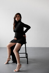 Pregnant woman in a black dress posing against a white wall, smiling and holding her belly.