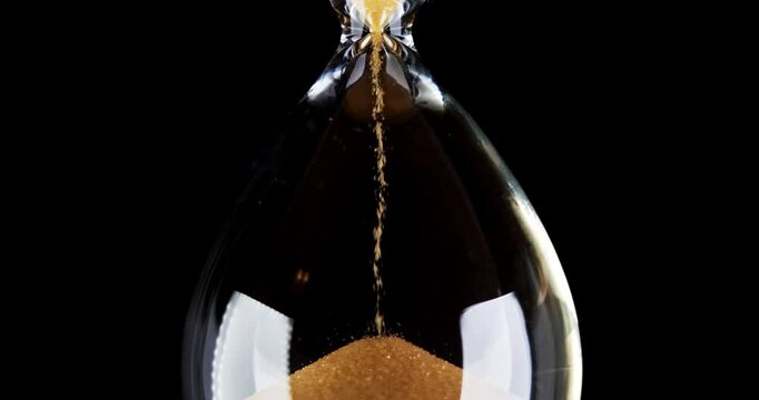 Hourglass. Sands move through hour glass. Sandglass close-up on a black background. Slow motion video. A pile of Golden sand at the bottom of the hourglass, small grains of sand fall from above.